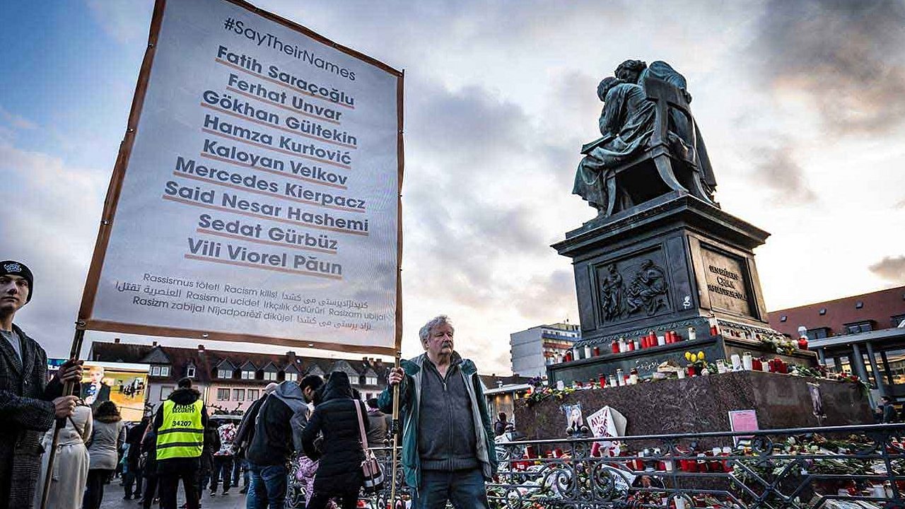 Plakat mit den Namen der Opfer aus Hanau, Blumen, Kränze und Kerzen am Brüder-Grimm-Denkmal auf dem Marktplatz 