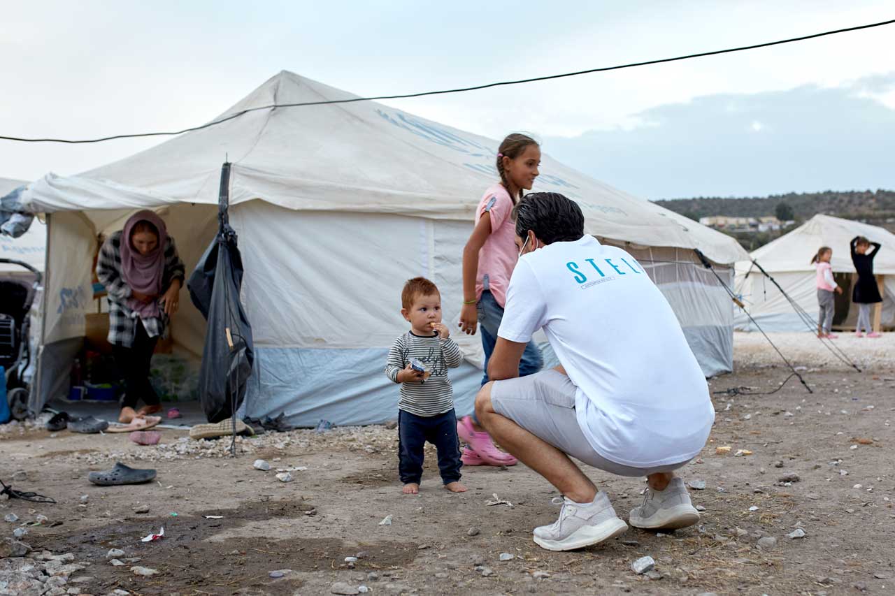 Serkan hockt vor einem Flüchtlingszelt. Vor ihm steht ein kleines Kind.