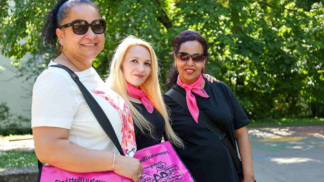Drei Frauen im Park mit pinken Halstüchern und pinken Taschen, auf denen "Stadtteilflüsterinnen" steht. Zwei von ihnen sind PoC