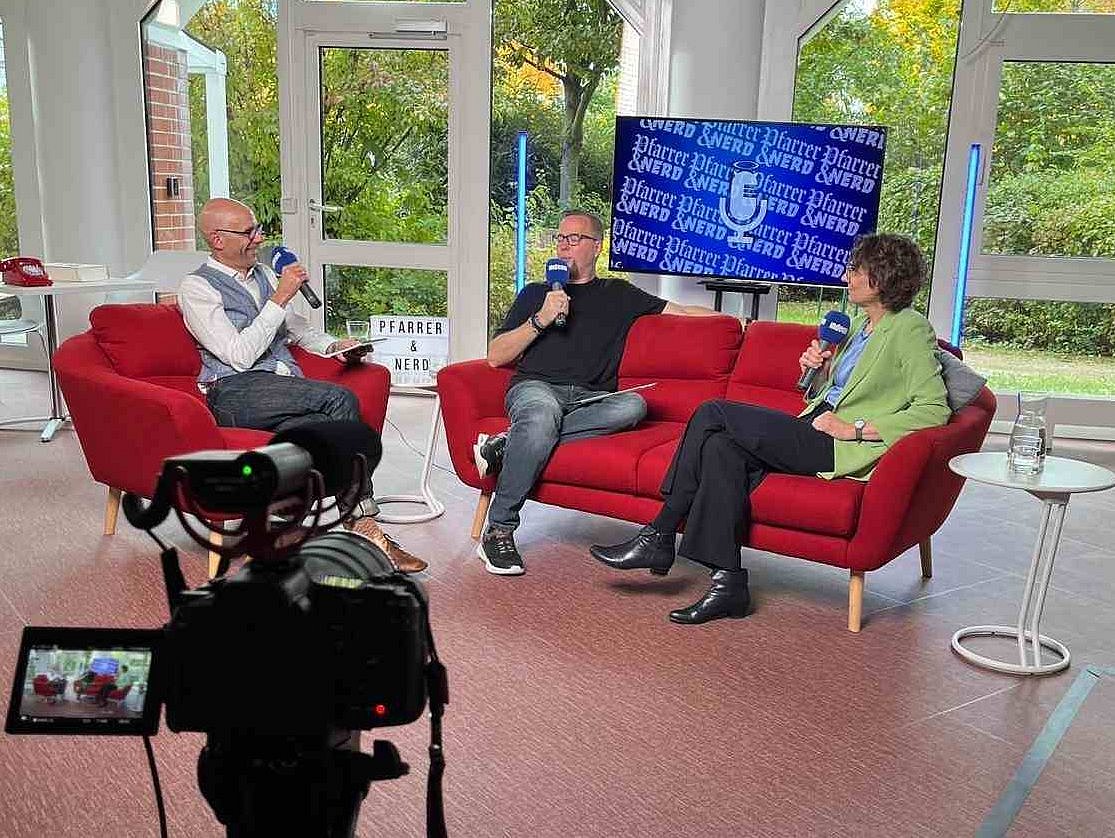 Blick auf eine Kamera und im Hintergrund sitzen Podcast-Host Martin und Seba mit der künfitgen Kirchenpräsidentin Christiane Tietz