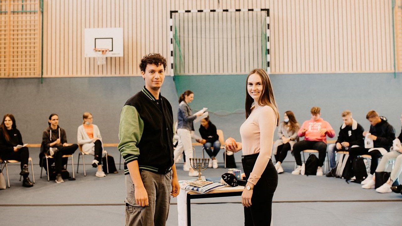 Zwei junge Juden und Jüdinnen stehen in einer Turnhalle vor einer Gruppe.