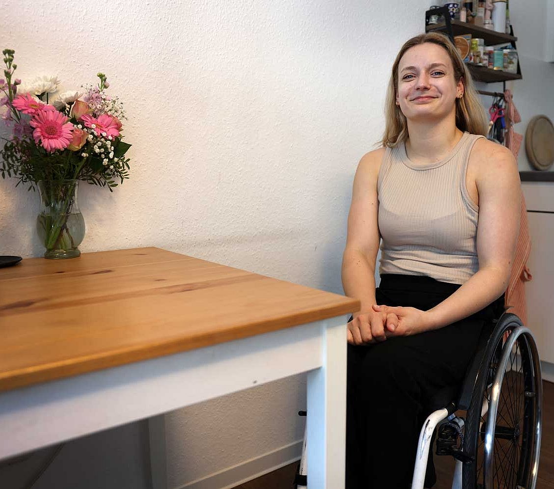 Catharina Weiß sitzt im Rollstuhl in ihrer Wohnung, im Hintergrund stehen drei Kerzen und ein rosafarbener Blumenstrauß