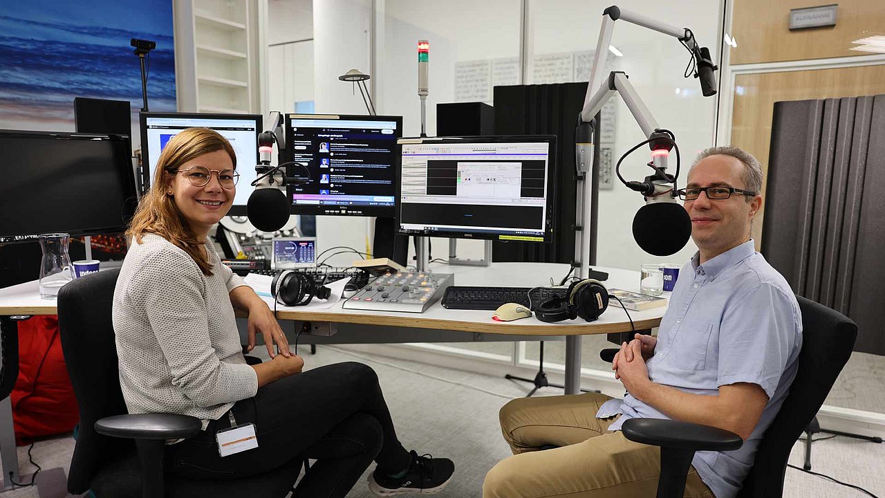 Charlotte Mattes und Jens Brehl lächeln in die Kamera. Sie sitzen im Studio. Im Hintergrund sieht man Bildschirme und Mikrofone. 