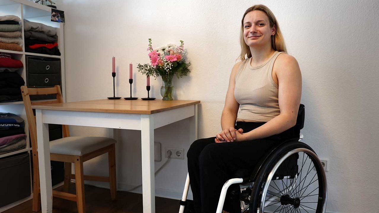 Catharina Weiß sitzt im Rollstuhl in ihrer Wohnung, im Hintergrund stehen drei Kerzen und ein rosafarbener Blumenstrauß