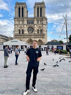 Simon steht auf einem Platz in Paris und dahinter ist Notre Dame zu sehen