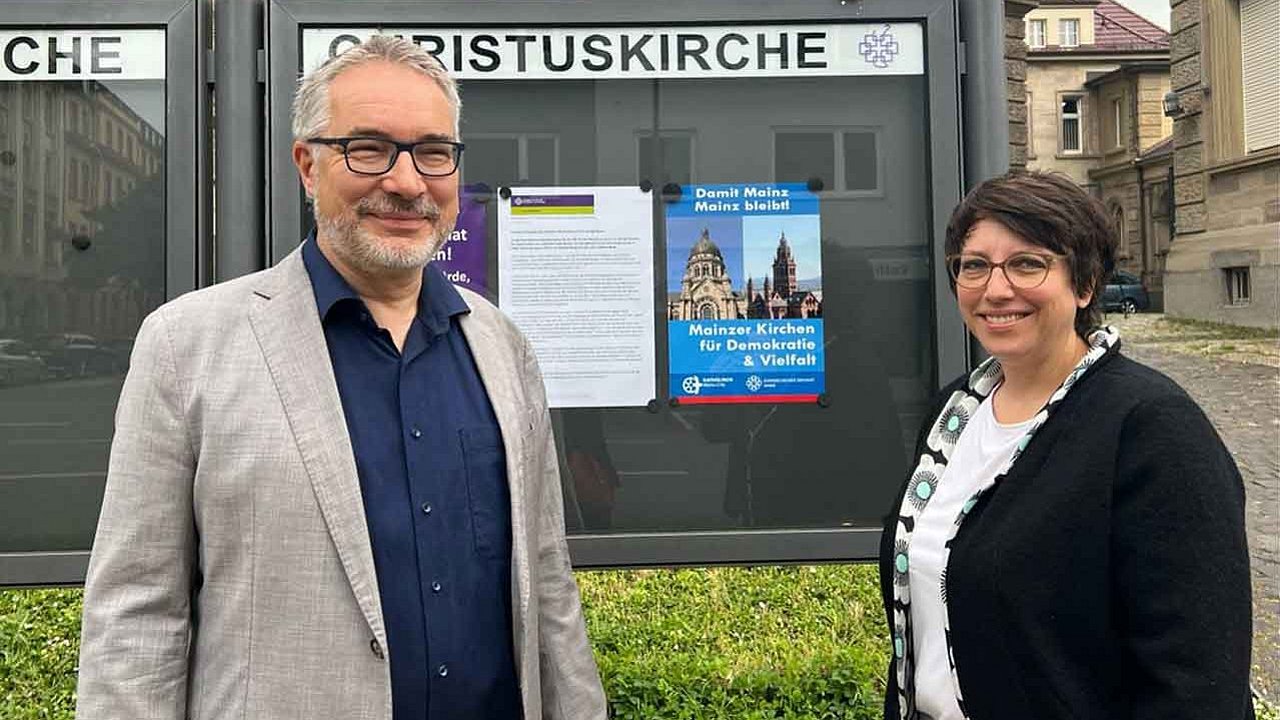 Pfarrerin Eva Lemaire und Dekan Andreas Klodt vor dem Schaukasten der Christuskirche mit der Plakataktion "Damit Mainz Mainz bleibt".