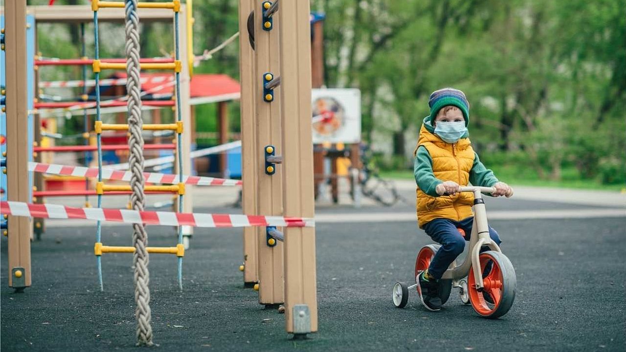 Ein Kind mit Fahrrad und Maske ist alleine auf einem Spielplatz. Die Spielgeräte sind abgesperrt