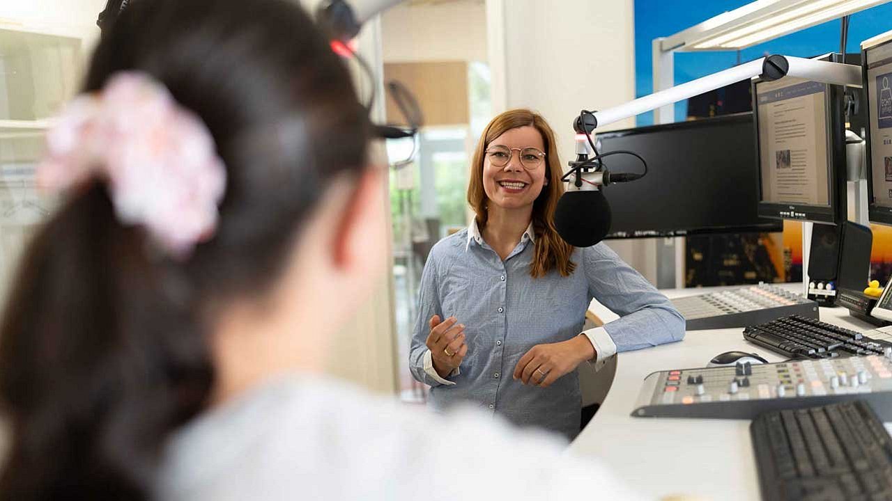 Im Vordergrund, unscharfer Pferdeschwanz, mit rosa Haargummi, im Hintergrund Host Charlotte Mattes, lächelt und gestikuliert im Aufnahmestudio.