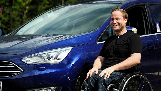 Lucas sitzt im schwarzen T-Shirt im Rollstuhl vor seinem blauen Ford C-Max