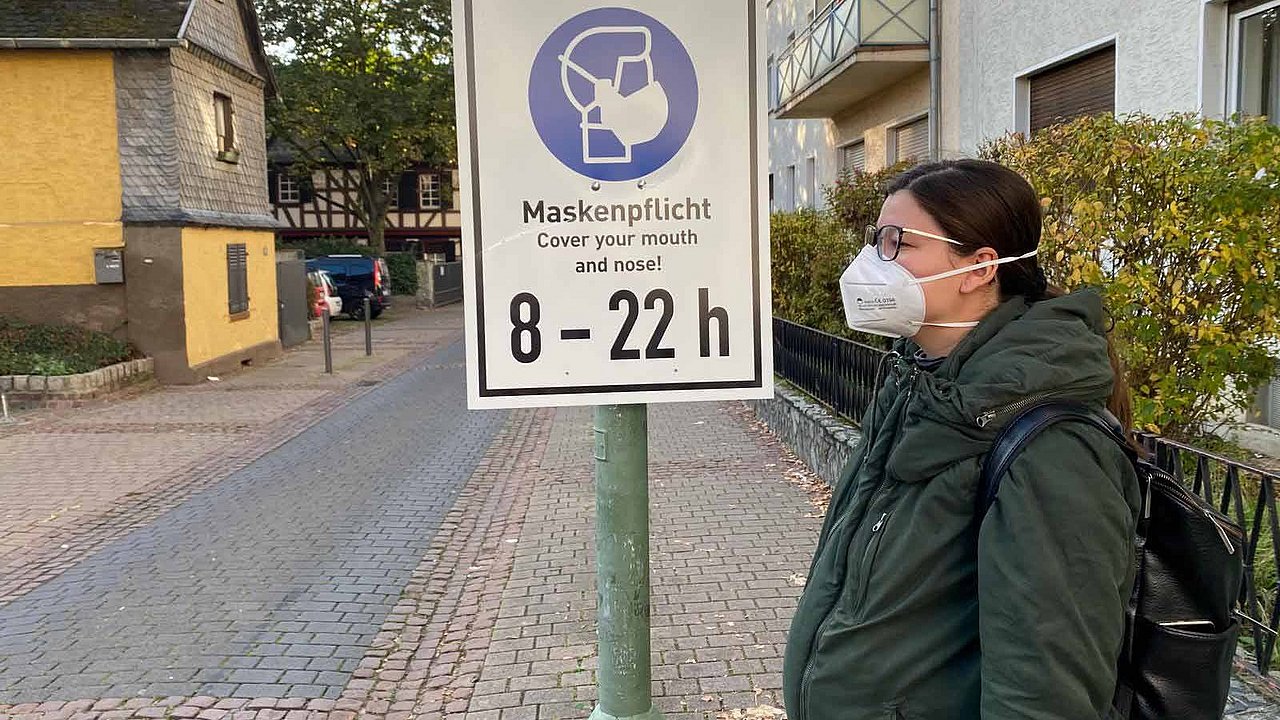 Charlotte Mattes, im Freien, mit Maske vor einem Schild, das an die Maskenpflicht erinnert.