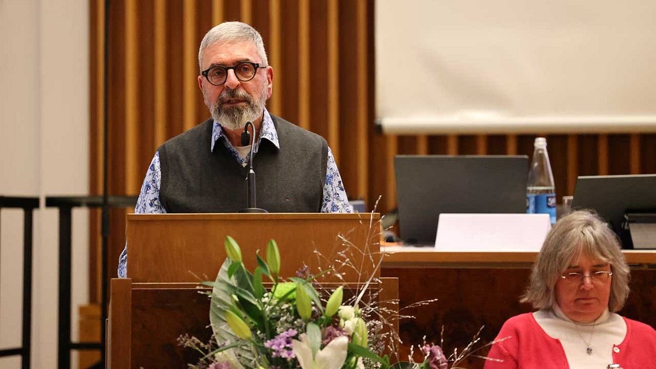 Matthias Schwarz steht am Rednerpult vor der Synode. Im Hintergrund sitzt Petra Knötzele.