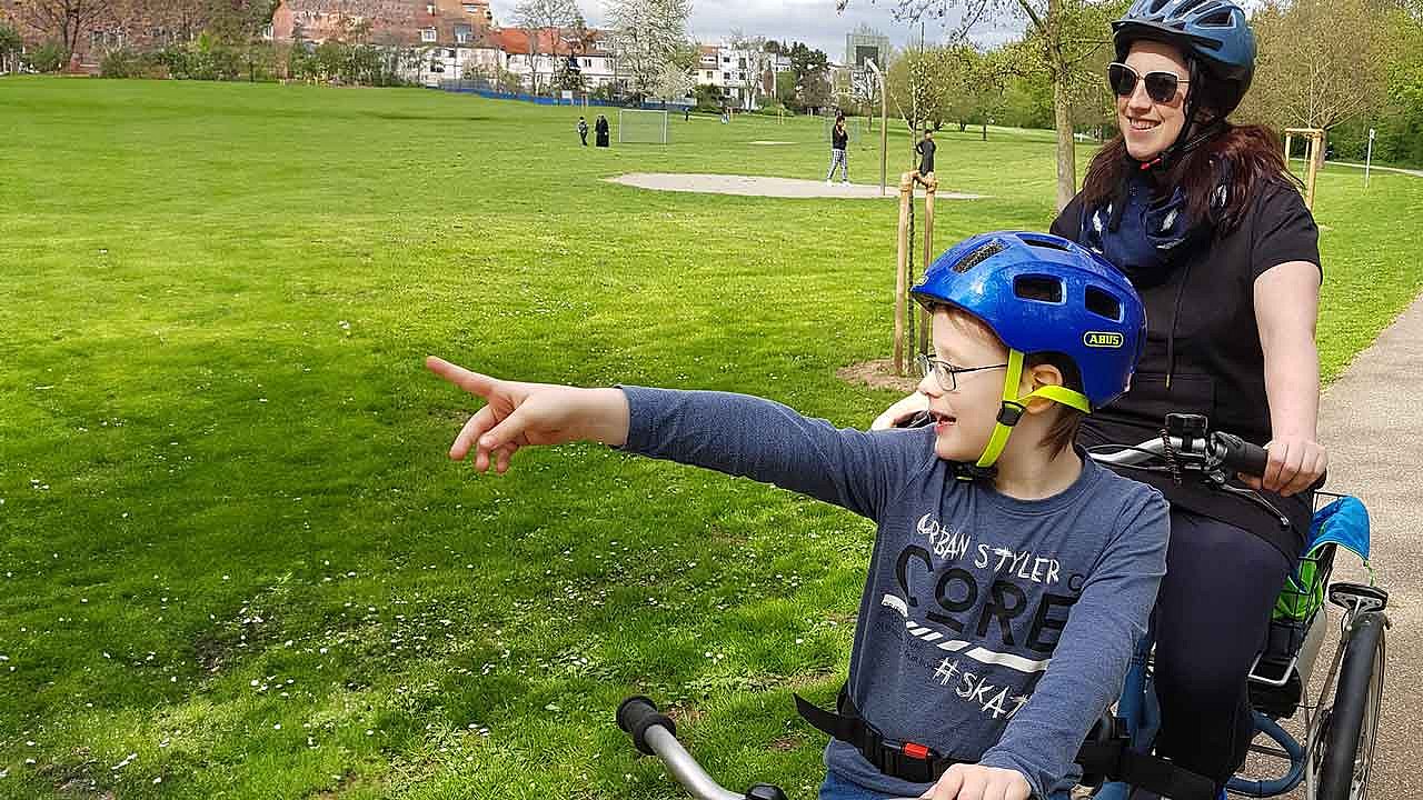 Lukas und seine Mutter Lisa fahren auf dem Therapietandem im Park an einer Wiese vorbei