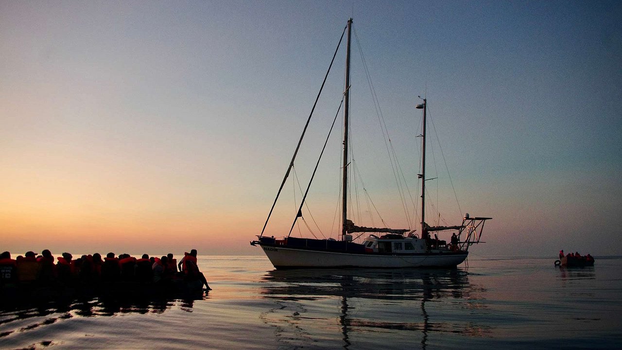 Segelschiff vor einem Schlauchboot mit vielen Geflüchteten darauf