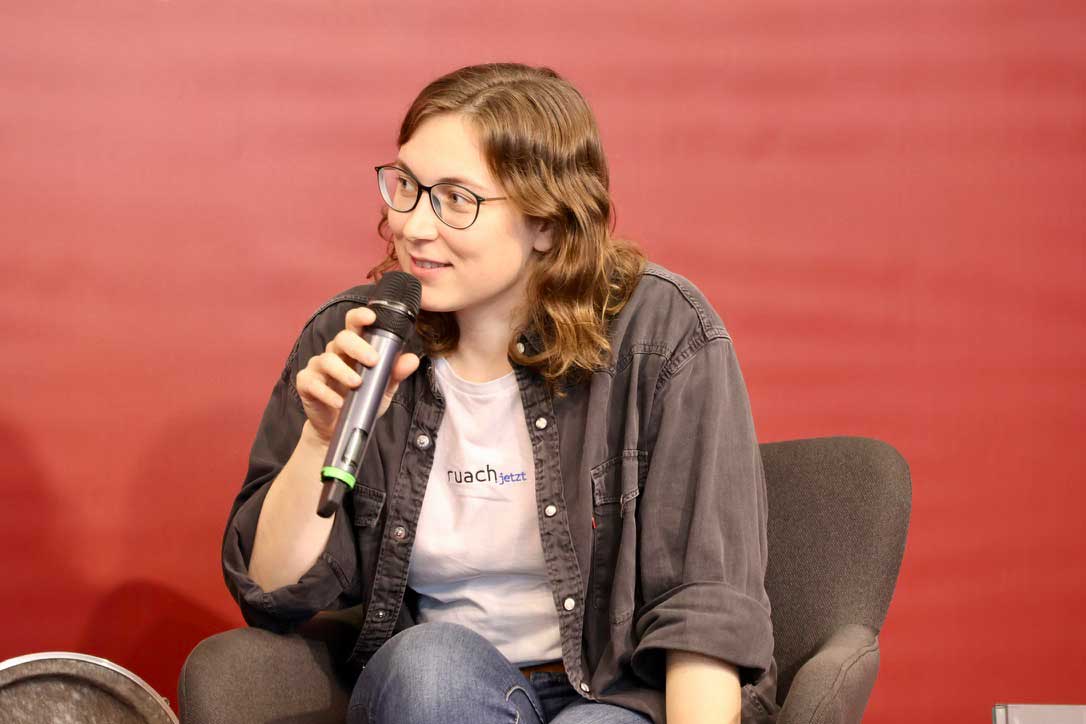 Lisa Menzel sitzt für ruach.jetzt auf einem Podium auf der Frankfurter Buchmesse 2023. Sie hält ein Mikrofon in der Hand und blinkt nach links. 