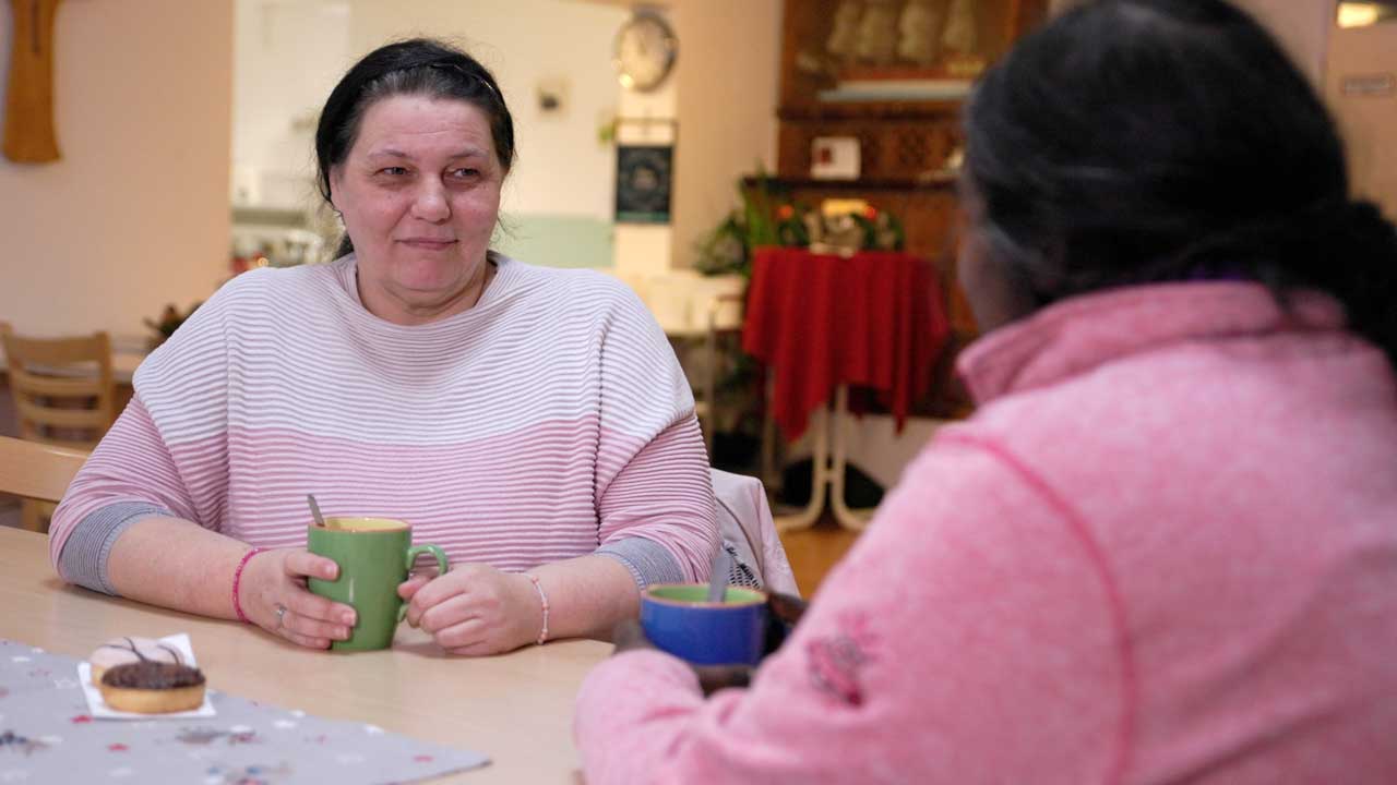 Cornelia sitzt am Tisch in der WG und blickt eine Mitbewohnerin an.