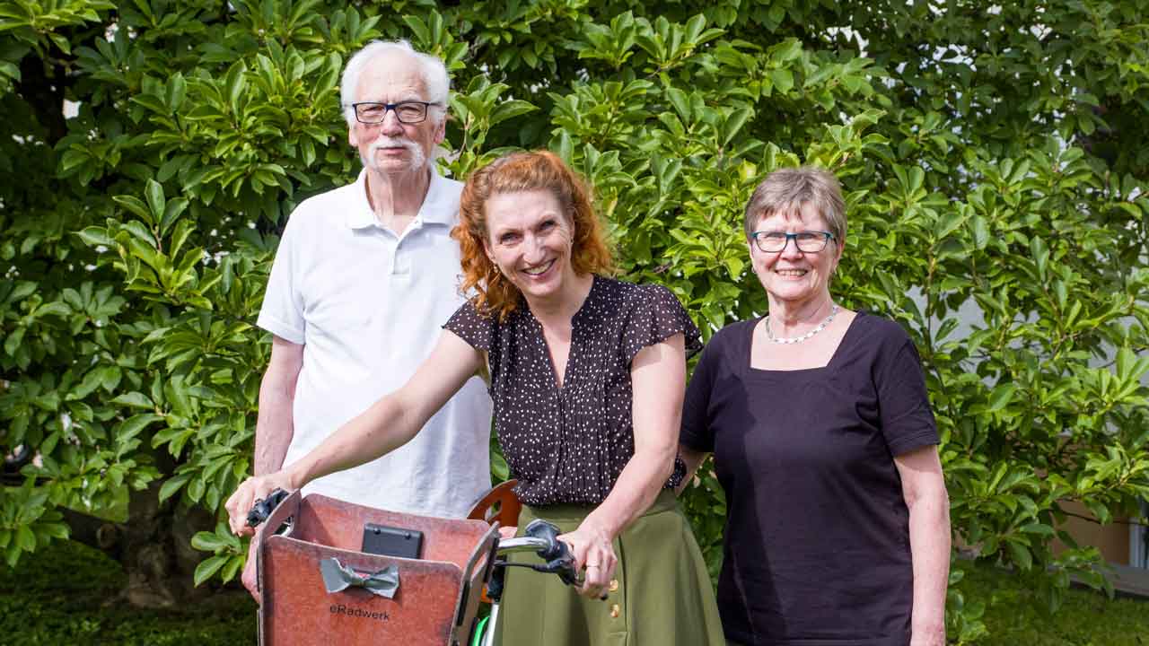 Janine und ihre Eltern stehen draußen. Janine hat ein Fahrrad in Händen. 