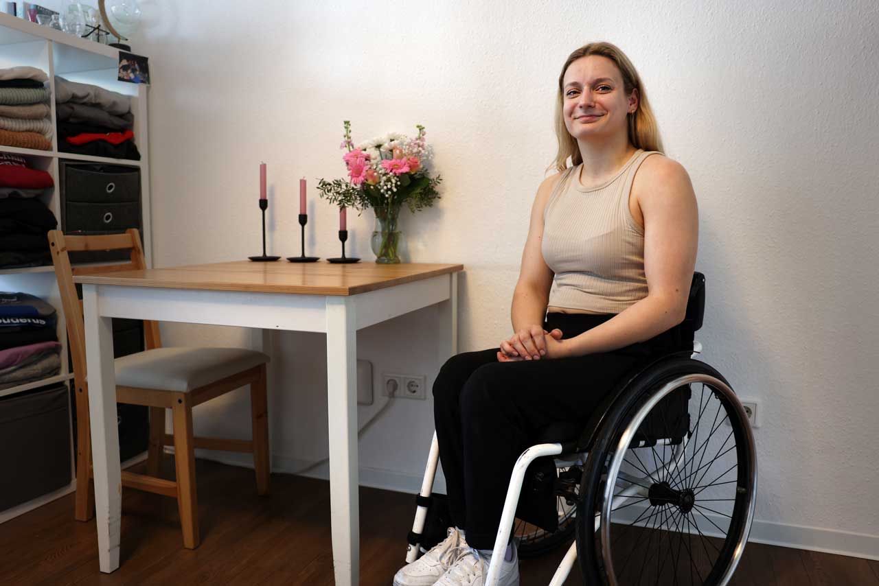 Catharina Weiß sitzt im Rollstuhl in ihrer Wohnung, im Hintergrund stehen drei Kerzen und ein rosafarbener Blumenstrauß