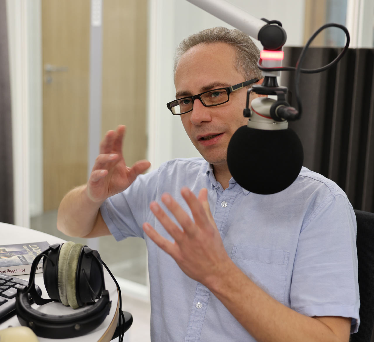 Jens Brehl, mit Brille, sitzt im Studio am Mikro, gestikuliert mit den Händen. 