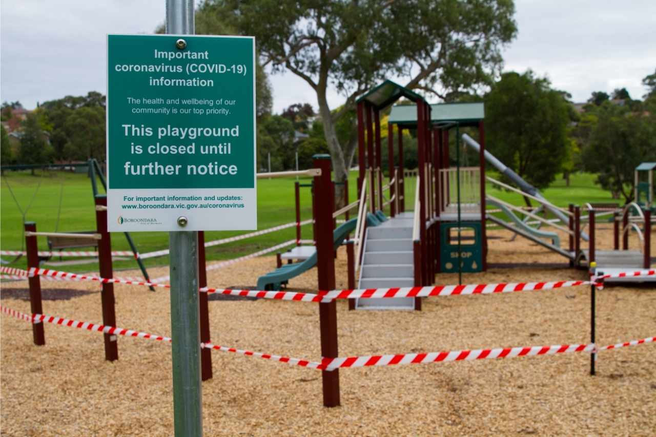 Ein leerer Spielplatz, abgesperrt mit weiß-rotem Flatterband. Vorne ein Schild, dass auf den Corona-Virus aufmerksam macht